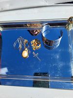 Mixed jewelry items arranged on reflective glass tray: necklace with white oval pendant, floral ring, cross pendant, wide cuff bracelet with etched design.
