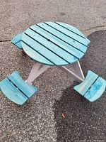 Photo of a small round kids picnic bench painted turquoise on table and seats, white legs, placed on asphalt surface.