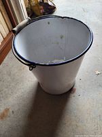 Top-down angle showing white metal bucket with black rim and wooden handle, placed on concrete floor.