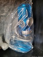 Blue microscope partially visible inside a transparent plastic bag, showing base, eyepiece and adjustment knobs.