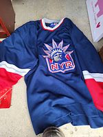 Full front view of navy blue New York Rangers hockey jersey with large embroidered logo and red, white stripes on sleeves.