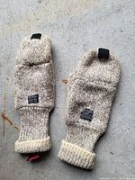 Photo of the pair of gloves laid flat showing the knitted wool exterior with folded mitten flap, black tab, and cuff.
