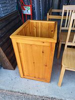 Photo of a solid wood rectangular box with an open top, natural light brown color, visible wood grain and knots, standing on a concrete floor among wooden chairs and other furniture.