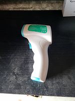 Side view of a white and turquoise handheld infrared temperature checker device resting on a black surface.