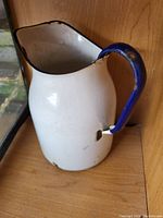 Full view of a white enamel water jug with a blue handle showing rust and wear.
