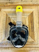Full face black and clear snorkel mask with yellow capped snorkel tube and adjustable black straps on wooden surface