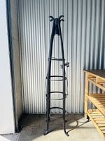 Full front view of the tall bedside lamp made of black metal, standing against a corrugated metal wall next to wooden shelves.