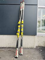 Pair of Skinetik 100xpi skis standing against a wall outdoors, showing silver and white color with red text and yellow bindings