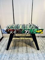 Front view of foosball table showing soccer-themed graphics on side panels and black angled legs.