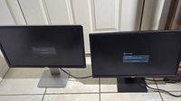 Two Dell 24-inch monitors side by side on tiled floor, both powered on showing settings menus, one with silver stand and one black stand.