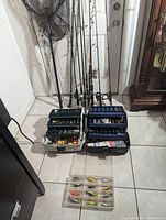 Photo showing six fishing rods standing against the wall with attached spinning reels, two open tackle boxes with compartments showing assorted terminal tackle and fishing accessories, and a transparent box with various fishing lures in front.