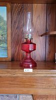 Front view of the complete antique red glass hurricane lamp placed on a wooden shelf.
