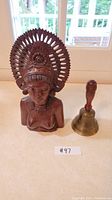 Full view of the wooden bust and brass bell side by side on a table with natural light.