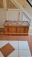 Front view of wooden storage bench showing carved floral panel and padded brocade cushion top.