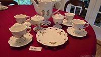 Full view of white bone china tea set on red tablecloth including teapot, cream, sugar, cookie plate, and four cups with saucers, all decorated with pink roses.