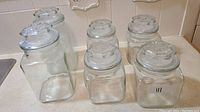 Photo showing six clear glass jars with matching lids arranged on a kitchen counter, two large and four medium-sized jars.