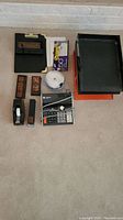 Overview of all office supplies including electric adding machine, letter trays, stapler, tape dispenser, stamps, clips, and clipboard.