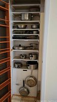 View of pantry shelves containing multiple stainless steel pots with lids, mixing bowls, colander, frying pans, baking dishes, and small kitchenware items.