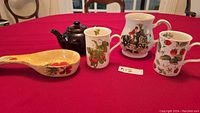 Shows large brown Betty teapot, Sadler hunt horse mug, two strawberry pattern china mugs, and ceramic spoon rack on red tablecloth.