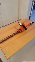 Bright orange Black & Decker electric hedge trimmer with 16-inch blade lying on a wooden table, clear view of blade and front handle.
