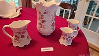 Photo of the entire set showing white bone china vase with pink rose design and three matching pitchers of varying sizes on red tablecloth.