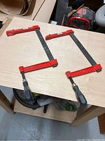 Two 12-inch sliding bar clamps lying on wood board, showing red metal bars and black plastic handles, used condition