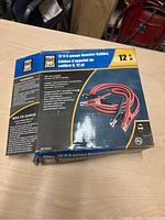 Box of 12 ft 6 gauge booster cables on a table, showing brand, length, and cable specifications.