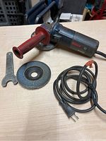 Bosch B9150 angle grinder on table with side handle attached, abrasive disc mounted, wrench beside it, and power cord coiled with plug.