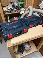 Two closed Bosch stackable carry cases side by side on a wooden table in a workshop, highlighting their size difference, blue color, red latches, and Bosch branding on the lids.