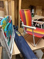 Three chairs shown: one red/orange/yellow striped folding beach chair, one blue/green floral striped folding beach chair, both with white metal frames; deluxe arm chair in blue carrying bag on table.