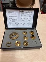 Open case showing six brass bushings and one brass adapter plate on foam insert with one empty slot.