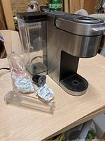 Front view of Keurig coffee machine showing water reservoir, coffee outlet, and several K-Cup pods and filters laid out beside it on table.
