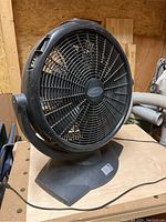 Front view of large 20-inch black rotary shop fan on stable base in workshop setting, showing blade grille and brand logo.