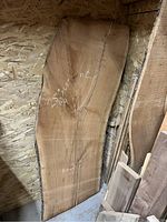 Large cherry wood slab leaning against plywood wall, showing natural wood grain and live edge, with some chalk markings on the surface