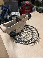 Extension cord reel showing beige plastic body, metal winding handle, and circular black net basket for cord storage on a wooden table with background tools.