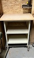 Front view of metal machine stand with wood top, two shelves, and rolling caster base in workshop setting.