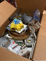 Box containing mixed electrical items including plugs, cords, remote control, and metallic electrical boxes