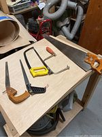 Multiple saws on plywood surface including Diston D8, coping saw, hacksaw, and keyhole saws.