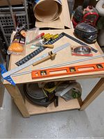 Top view of a work table displaying various tools and shop items including rulers, screwdrivers, safety glasses, sanding block, braided rope, and spirit level arranged on its surface.