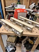 Three beige plastic tip-out bin organizers with six clear drawers each placed on a wooden peg board workbench; workshop environment visible in background.
