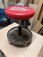 Photo shows a red cushioned round seat with white Big Red logo, height adjustment lever, chrome adjustable column, black circular base with dirt and five orange caster wheels.