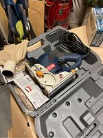Ryobi biscuit jointer placed in its grey molded plastic case with power cord coiled, alongside the dust collection bag and a small packet of biscuits visible inside the case.