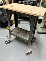 Photo shows a small metal frame machine stand with a rectangular plywood top fixed by bolts, four legs with locking caster wheels, and a lower metal shelf with vent holes. Metal frame surface shows signs of use and requires cleaning.