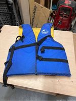 Blue and yellow universal size life jacket with black adjustable straps laid flat on wooden surface.