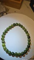 Photo showing the round green jade beads bracelet laid flat on a white circular surface. Beads show slight color variation from light to dark green.