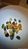 Multiple small natural mixed color tourmaline crystals displayed on a white surface under light showing range of greens, yellows, oranges, and browns.