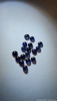 20 small natural calibrated blue sapphire round cabochons shown spread out on a white background under directional lighting. Stones display deep blue color with visible inclusions and translucency.