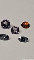 Photo showing five loose faceted gemstones arranged on white background including orange garnet, pink amethyst, and three blue/purple tanzanite stones.
