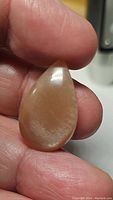 Close-up photo of the pear-shaped peach moonstone cabochon held between fingers, showing the smooth polished surface and peach color with shimmer effect.