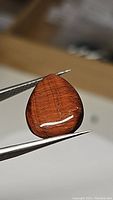 Natural red tiger's eye pear-shaped cabochon gemstone held by tweezers showing polished smooth surface and reddish-brown color
